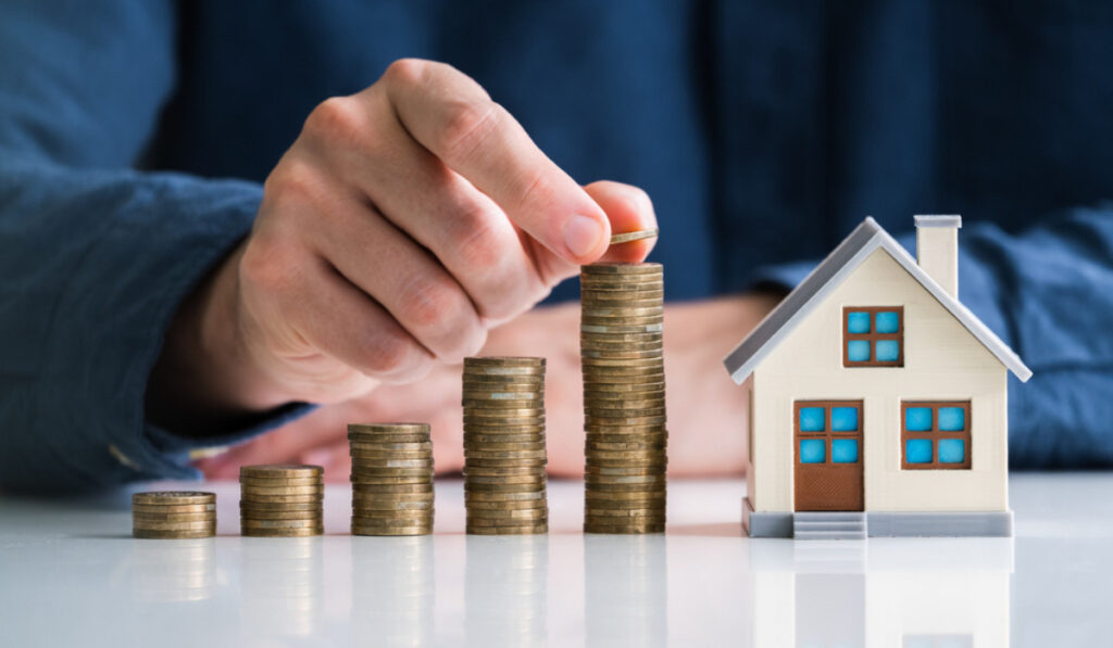 How to Invest in Real Estate with No Money: Proven Strategies for 2026 Hand stacking coins beside a house model symbolizing investing in real estate with no money and building passive income